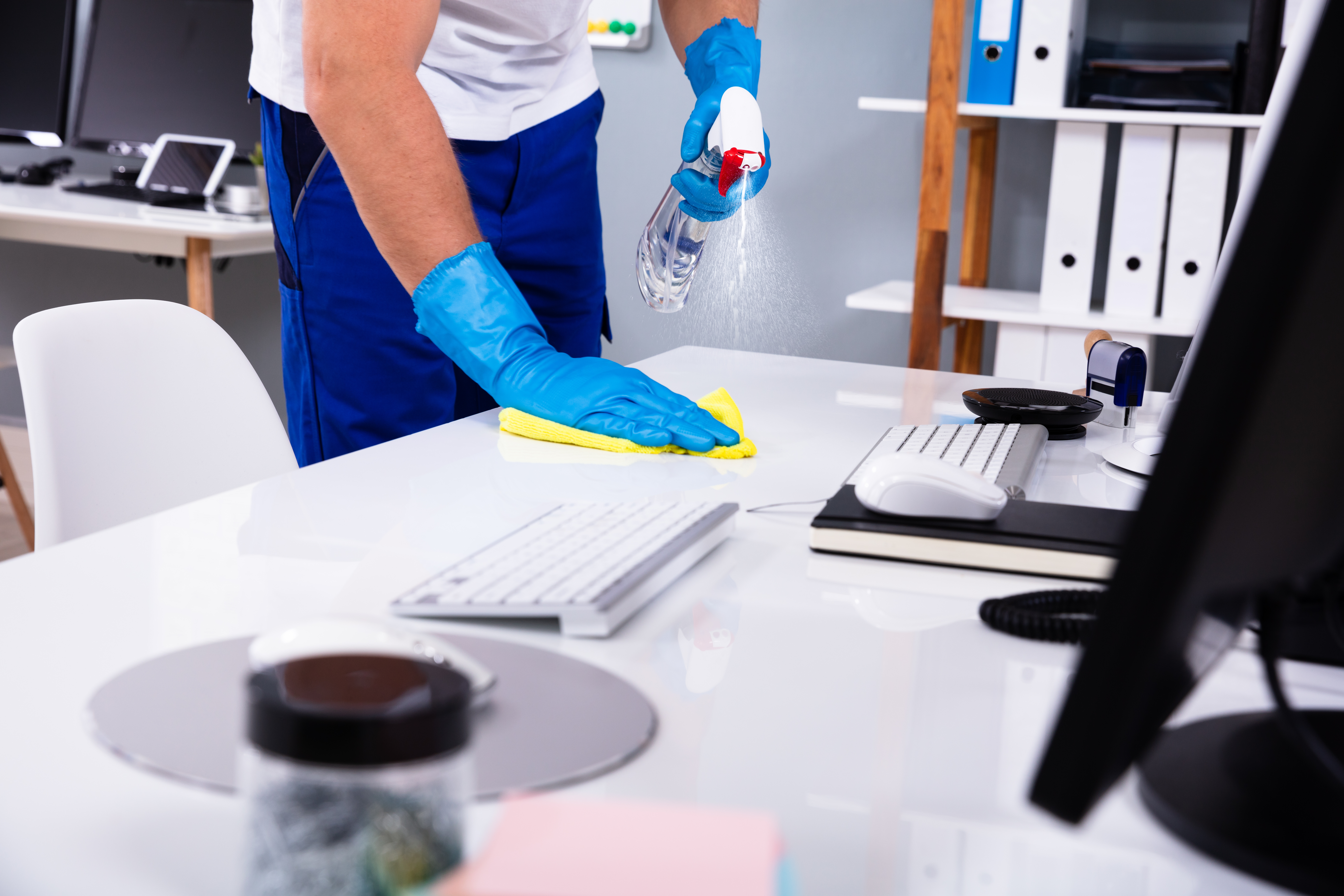 Een schoonmaker in een kantooromgeving maakt een wit bureau schoon met een doek en schoonmaakmiddel.